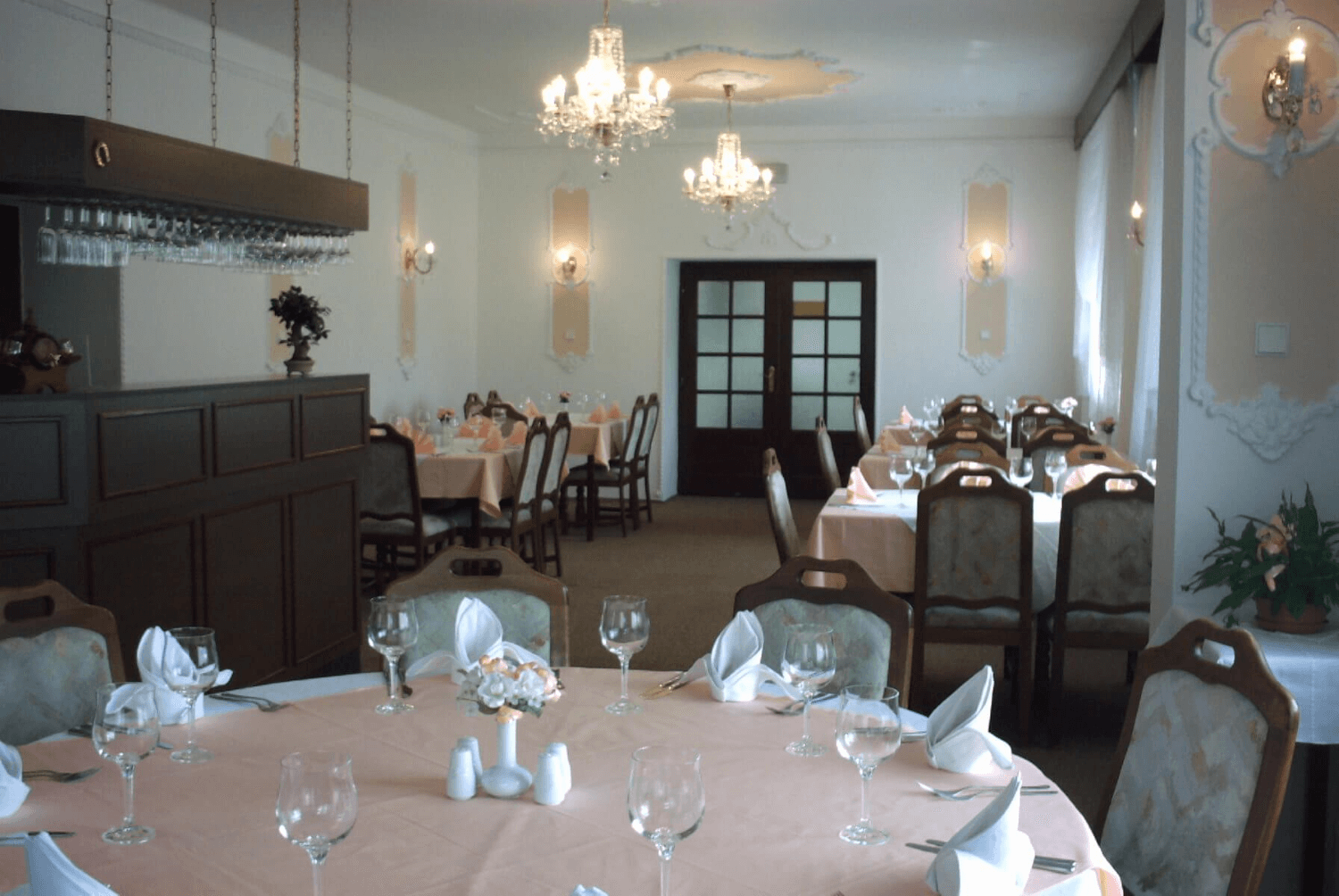 Jídelna hotelu, v popředí velký kulatý stůl pro více osob, v pozadí několik dalších stolů a vchod do jídelny.